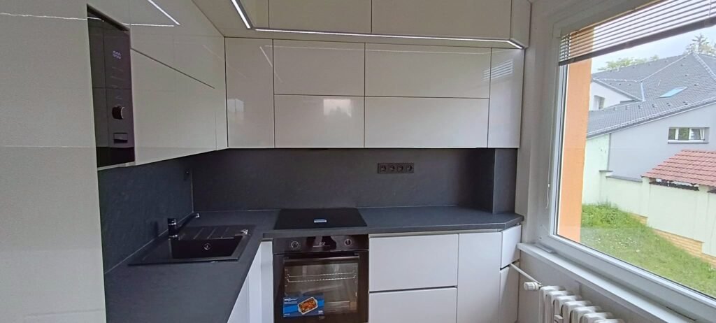 Bílá lesklá kuchyně s tmavým obkladem a oknem